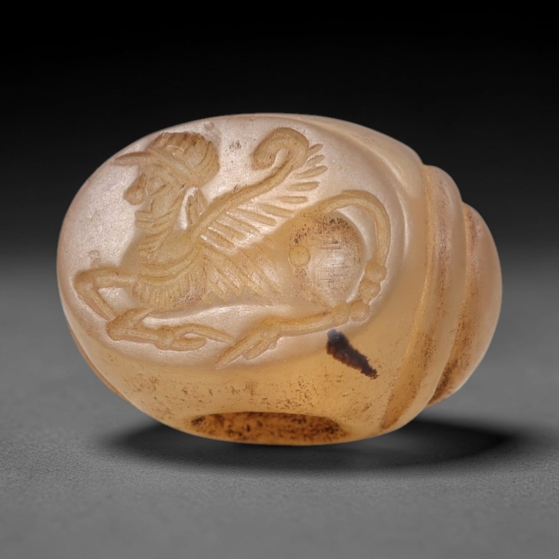 Ancient Chalcedony Hemispherical Stamp Seal - Near Eastern Winged Creature Amulet, 24AN: This ancient Near Eastern hemispherical stamp seal is carved from translucent, warm amber chalcedony, with a smooth, polished surface and natural dark inclusions. The seal's oval face depicts a winged