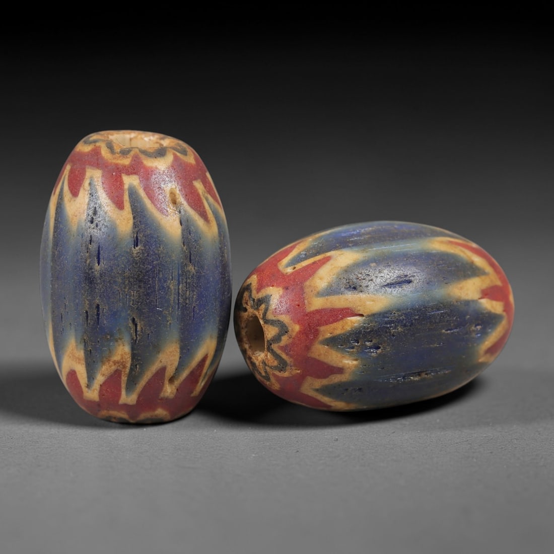 A Pair of Venetian Glass Chevron Beads, 18th-19th Century, 74AC: Crafted from translucent glass with a pale yellow core, each bead is decorated with a chevron pattern of alternating deep blue and vibrant red layers, radiating from the central perforation to the out