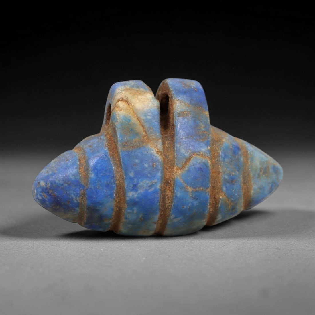 Ancient Lapis Lazuli Amulet - Egyptian Shell Form Pendant, 18AP: Carved from lapis lazuli, this amulet features a symmetrical, shell-like form with two raised upper lugs pierced for suspension, its body incised with horizontal bands that mimic the segmented structu