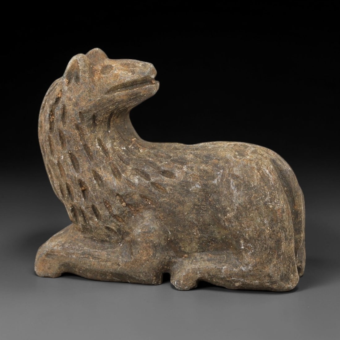 Ancient Stone Recumbent Feline Amulet - Near Eastern Lion Effigy, 9AM: This amulet is carved from textured grey-brown stone, depicting a stylized feline (lion or big cat) in a recumbent, crouched posture. The piece features incised linear and stippled detailing defining