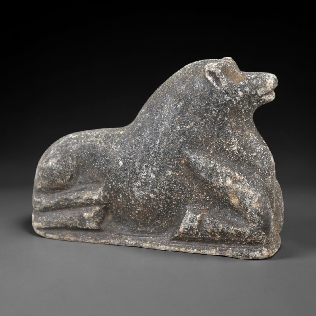 Ancient Stone Canine Amulet - Near Eastern Dog/Wolf Figurine, 45AM: This amulet is carved from dark speckled stone, depicting a stylized canine (dog or wolf) in a recumbent, crouched posture. The piece features incised linear detailing defining the facial features, ea