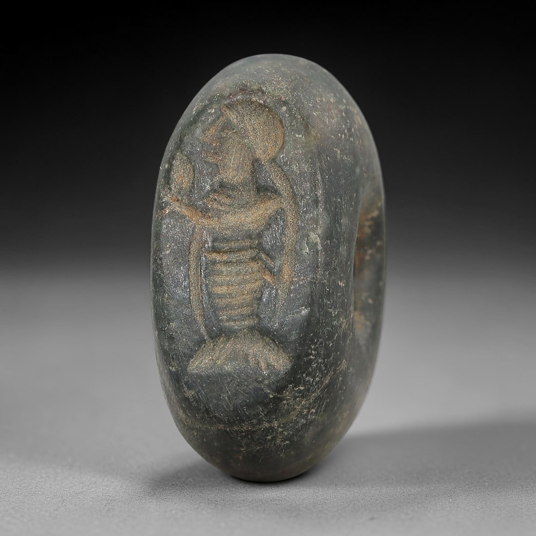 Ancient Stone Hemispherical Stamp Seal - Near Eastern Anthropomorphic Figure, 79AN: Carved from a dense, dark grey stone, this hemispherical stamp seal features a flat, oval face incised with a standing anthropomorphic figure. The figure is depicted with a headdress, a pleated garmen