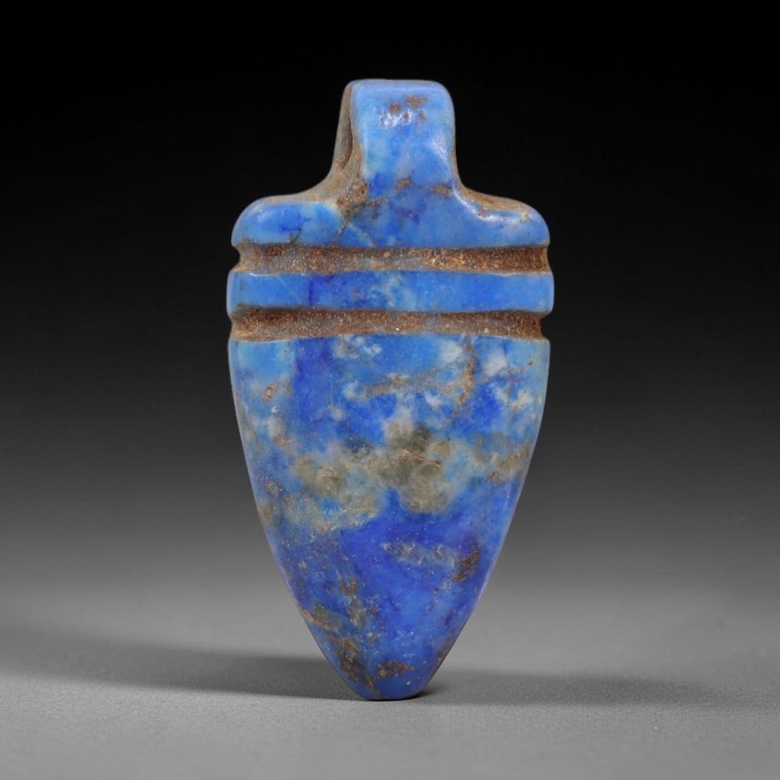 Ancient Lapis Lazuli Amulet - Egyptian Teardrop Pendant, 12AP: Carved from lapis lazuli, this amulet features a tapering teardrop form with a broad, flattened upper lug pierced for suspension, its body marked by two incised horizontal bands that define its struct