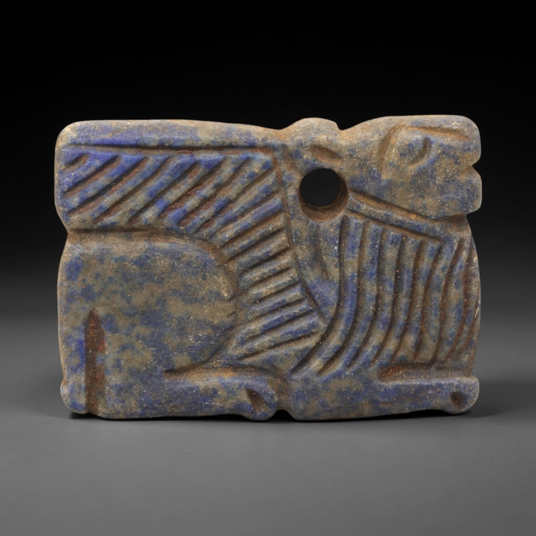 Ancient Lapis Caprid Amulet - Near Eastern Figurine, 49XV: Carved from lapis lazuli, this amulet depicts a stylized caprid (goat or sheep) in a recumbent posture, with incised linear striations defining the body and facial features, and a distinct perforation