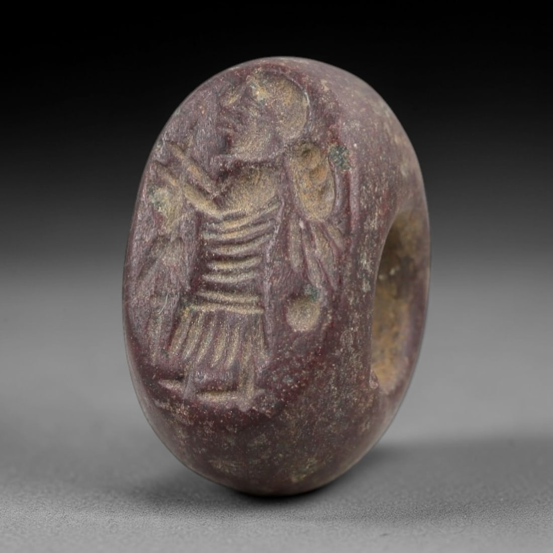 Ancient Stone Hemispherical Stamp Seal - Near Eastern Anthropomorphic Figure, 40AN: Carved from a dense, dark reddish-brown stone, this hemispherical stamp seal features a flat, oval face incised with a standing anthropomorphic figure. The figure is depicted with a headdress, a garme