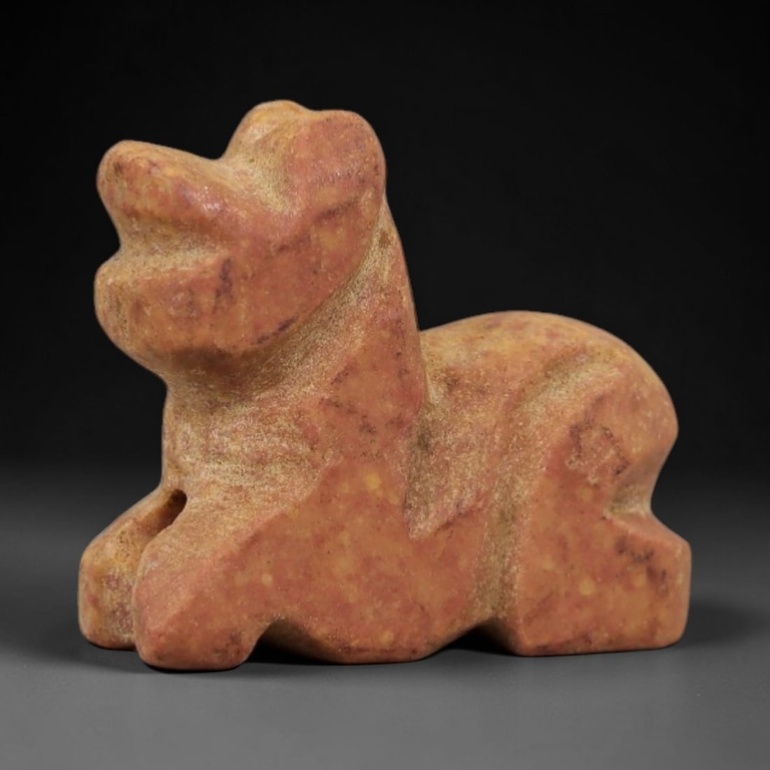 Ancient Reddish Stone Recumbent Feline - Near Eastern Amulet, 86AD: Carved from warm reddish-tan stone with natural granular texture, this amulet depicts a recumbent feline in a compact, stylized form. The creature is rendered in a prone resting posture, with simplifi