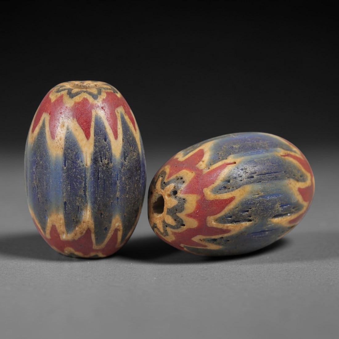 A Pair of Venetian Glass Chevron Beads, 18th-19th Century, 81AC: Crafted from translucent glass with a pale yellow core, each bead is decorated with a chevron pattern of alternating deep blue and vibrant red layers, radiating from the central perforation to the out