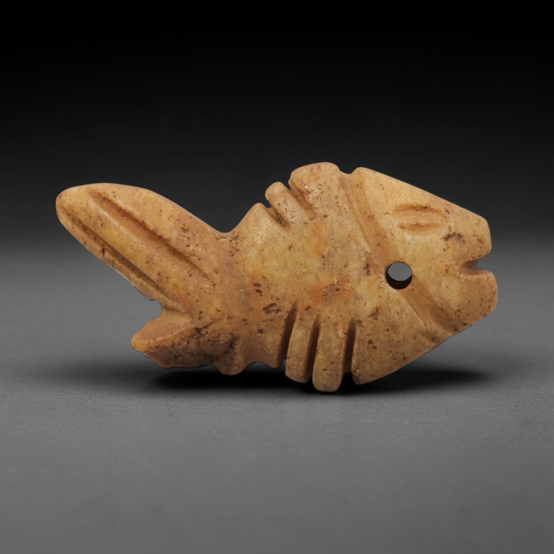Ancient Stone Fish - Egyptian Amulet Pendant, 60XR: Carved from pale orange stone, this amulet depicts a stylized fish with incised linear details representing fins and a drilled hole for suspension. The form and iconography are characteristic of Ancie