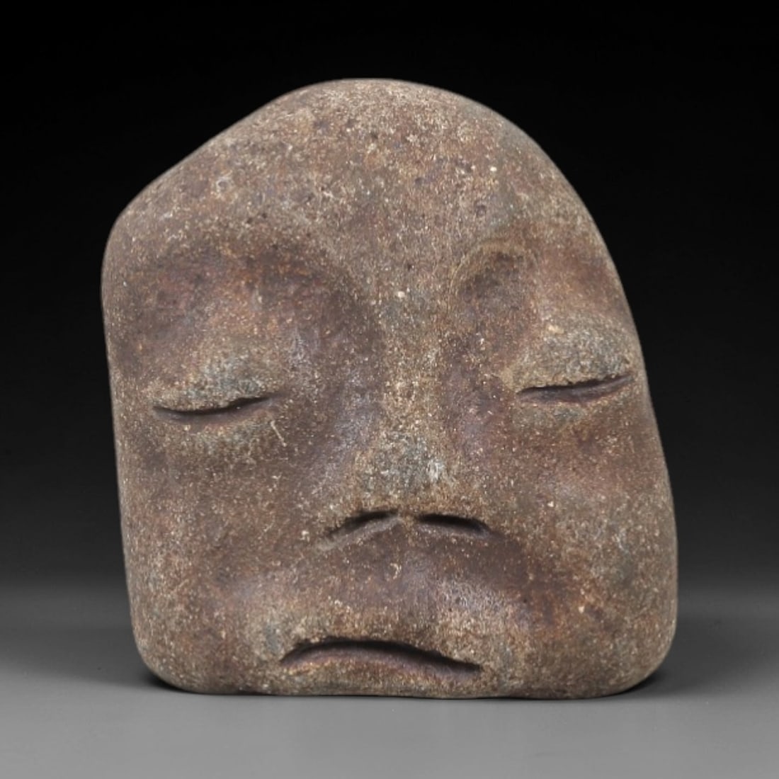 Ancient Stone Maskette - Near Eastern Amulet Figurine, 11FX: Carved from rough, granular stone, this small maskette features a stylized human face with closed, slanted eyes, a split nasal opening, and a downturned mouth, creating a serene, contemplative express
