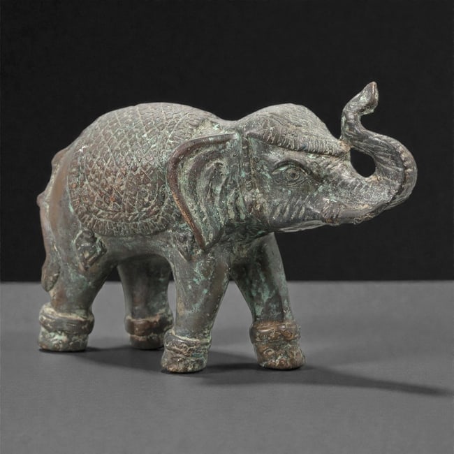 A Southeast Asian Bronze Elephant Figure, 20th Century, 20FJ: Cast in bronze, the figure represents a standing elephant shown in profile with the trunk raised and gently curved. The body is modeled with simplified but clearly articulated anatomical features, inc