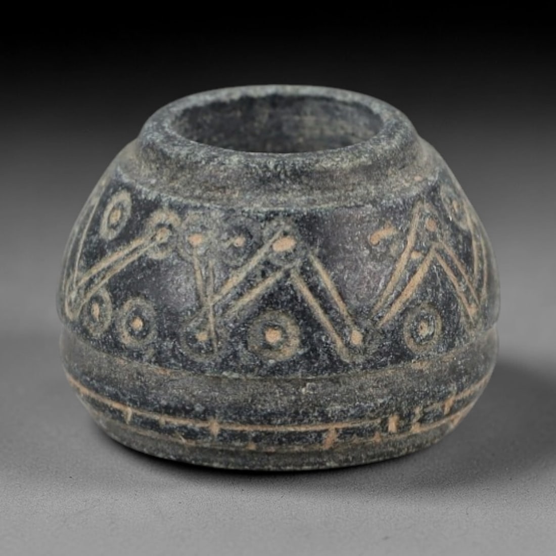 Ancient Stone Spindle Whorl - Egyptian Amulet Relic, 85XR: This artifact is carved from dark stone, exhibiting a compact spindle whorl form with a central perforation intended for suspension. The exterior surface is adorned with incised geometric motifs inclu