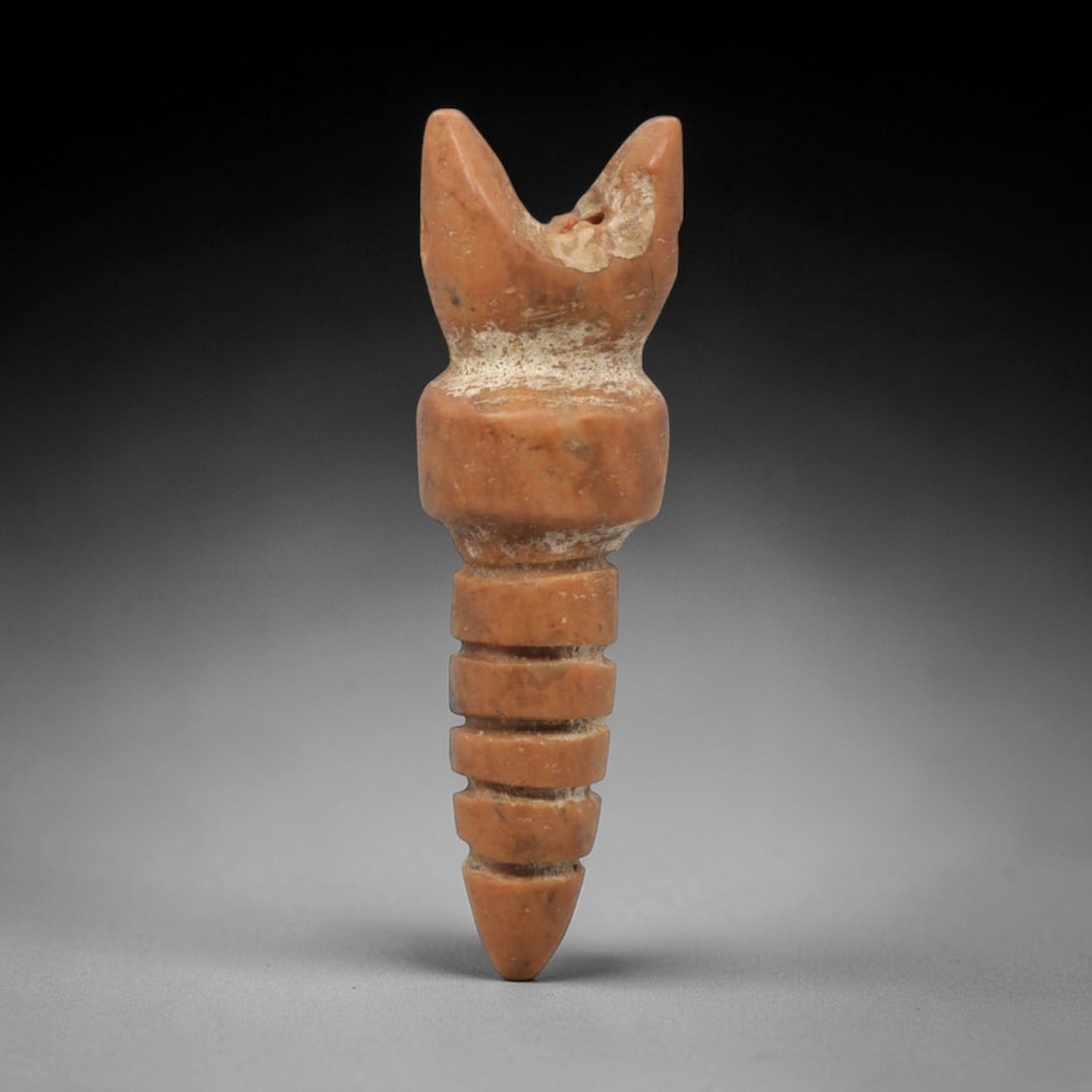 Ancient Red Stone Amulet - Near Eastern Ritual Figure, 59ZW: This amulet is carved from a rich red stone, featuring a stylized upper form with a V-shaped notch at the apex, surmounting a segmented, tapering lower body with horizontal incised rings. The form sug