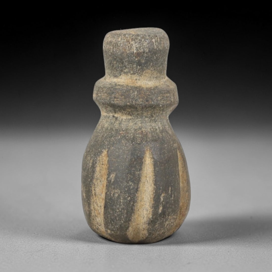 Ancient Stone Amulet - Egyptian Carved Pendant, 68AR: This artifact is carved from a dark, granular stone with lighter, striated mineral inclusions, exhibiting a weathered, matte surface consistent with age. The form is a compact, bottle-shaped amulet wi