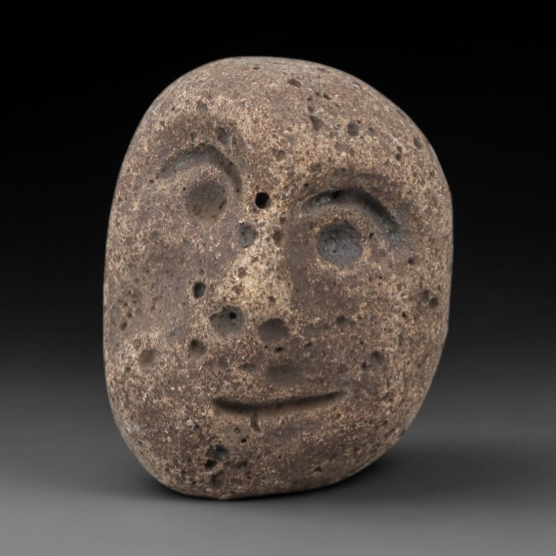 Ancient Volcanic Stone Maskette - Near Eastern Amulet Figurine, 20FX: Carved from porous volcanic stone, this small maskette features a stylized human face with incised eyes, a perforated nasal opening, and a horizontal mouth line, creating a gentle, smiling expression.