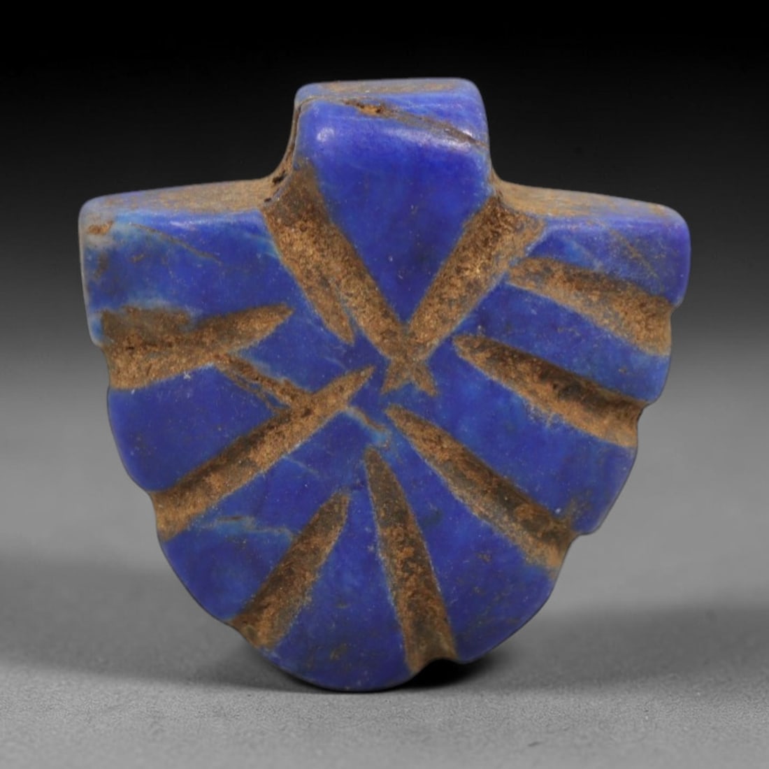 Ancient Lapis Lazuli Amulet - Egyptian Symbolic Pendant, 26ZT: Carved from lapis lazuli, this amulet features a shield-like form with a raised central boss pierced for suspension, its surface incised with radiating linear details that define symbolic protective m