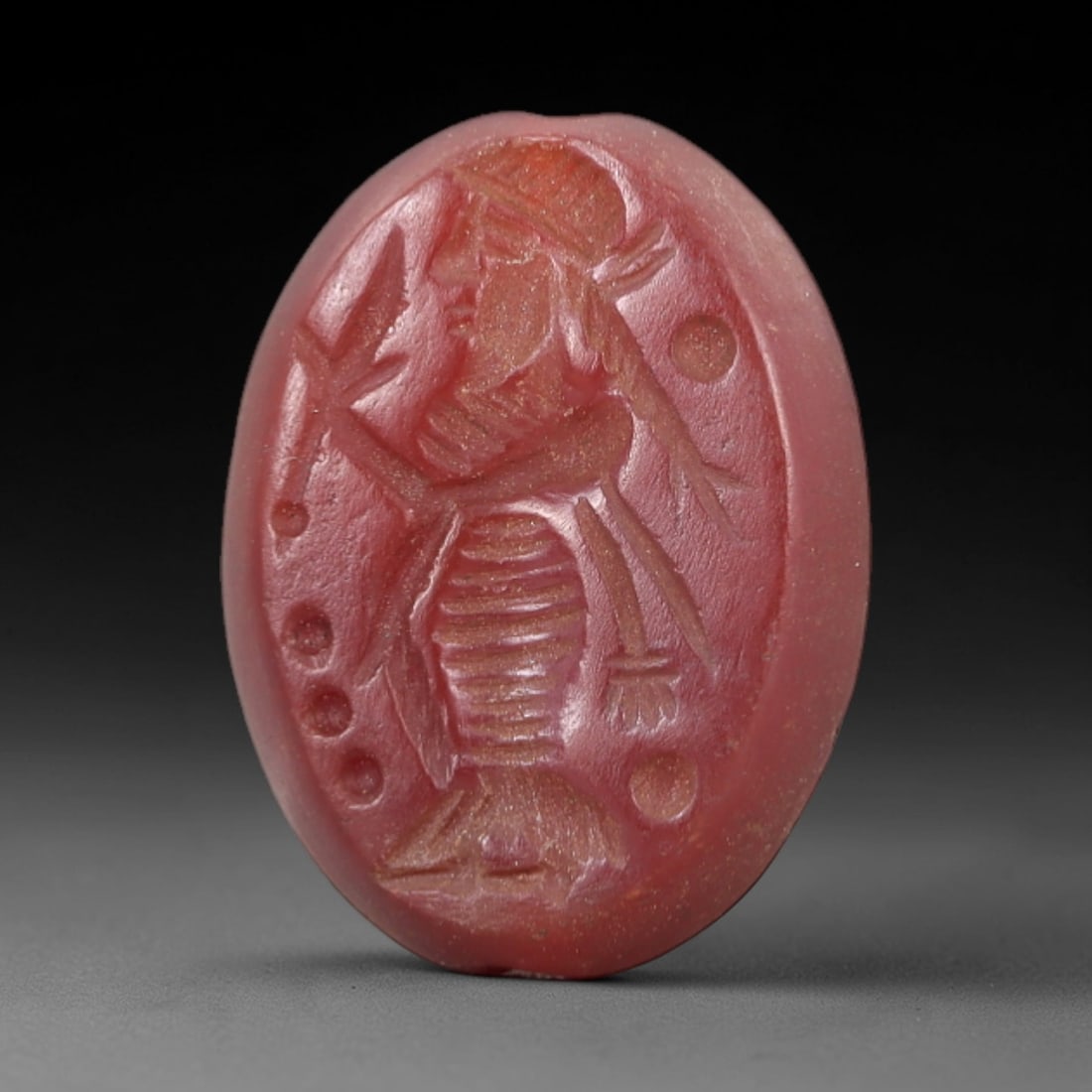 Ancient Carnelian Intaglio - Near Eastern Deity Figure, 19AL: Crafted from carnelian, this oval intaglio bears a deeply incised depiction of a robed, bearded figure holding a plant-like attribute, surrounded by circular dot motifs. Attributed to ancient Near Eas