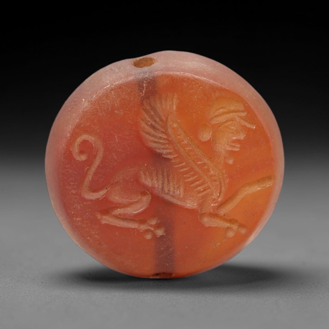Ancient Carnelian Intaglio - Sphinx Figure Seal, 37XV: Crafted from polished banded carnelian, this circular intaglio seal depicts a striding sphinx-a winged lion with a human head-adorned with a stylized cap and earring. The creature's feathered wings, s