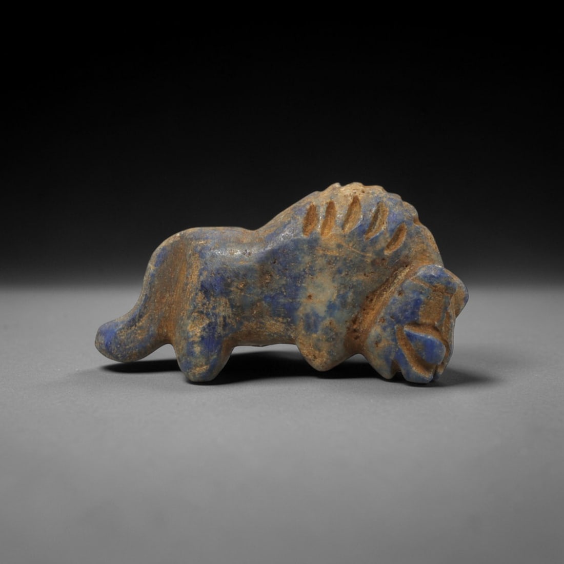 Ancient Lapis Lazuli Lion - Near Eastern Amulet Figurine, 14XT: Carved from lapis lazuli, this amulet depicts a striding lion in a stylized, compact form. The head is prominent with incised eye motifs and a defined muzzle, while the body features bold, vertical gr