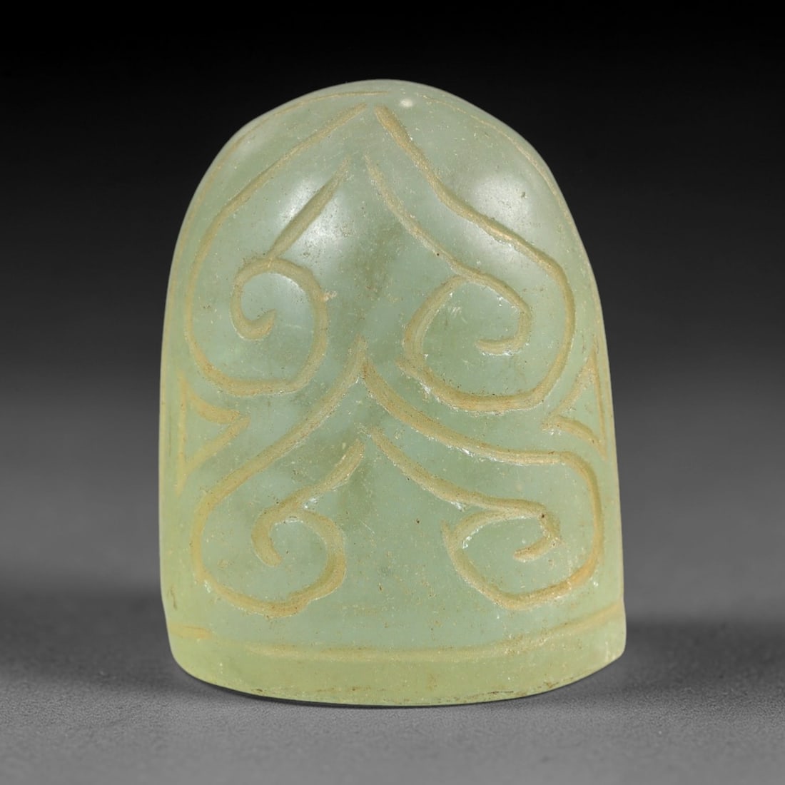 Ancient Glass Medieval Relic - Near Eastern Carving, 69AL: This artifact is crafted from translucent pale green ancient glass, formed into a domed, cylindrical shape with incised curvilinear decorative motifs including symmetrical scroll patterns. The carving