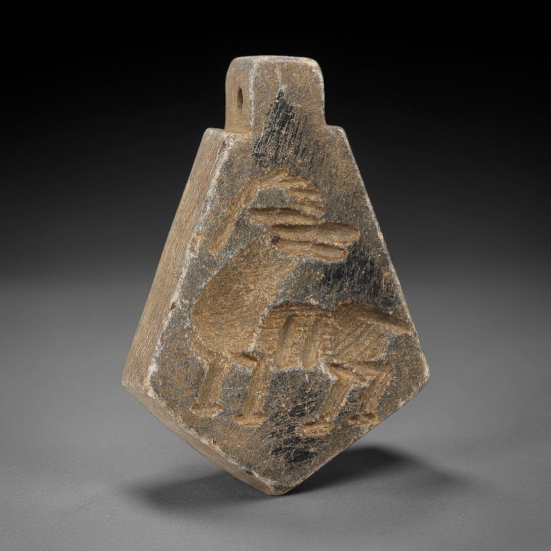 Ancient Schist Amulet Pendant - Ancient Relic, 96AG: Carved from schist, this ancient amulet pendant features a stylized animal form with subtle linear detailing typical of ancient artistic traditions. The stone exhibits a muted earthy hue with natural