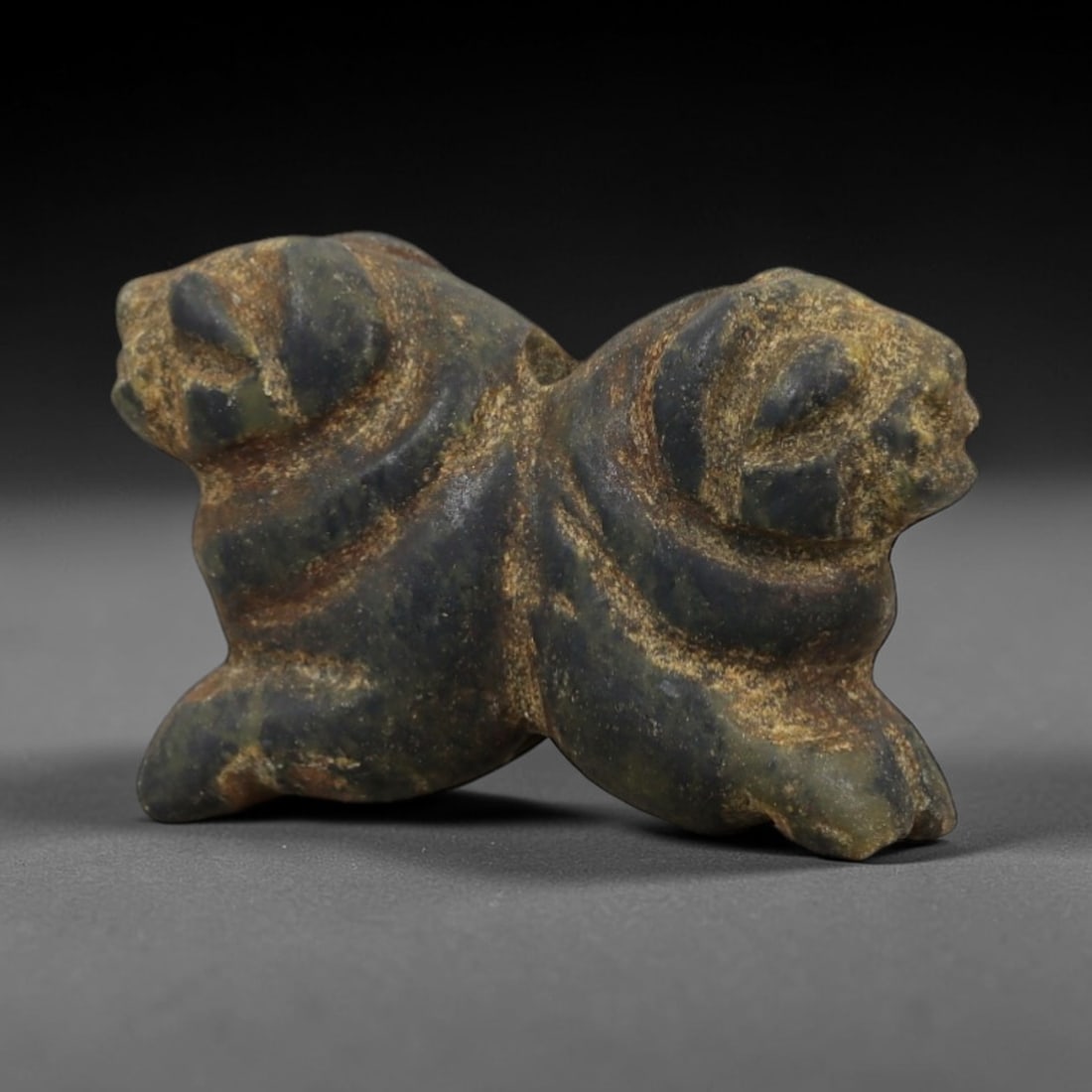 Ancient Dark Stone Double Zoomorphic Amulet - Near Eastern Relic, 92AD: Carved from dark greenish-black stone with tan mineral patination, this amulet features a stylized double zoomorphic form, depicting two contiguous animal heads with rounded contours and incised facia