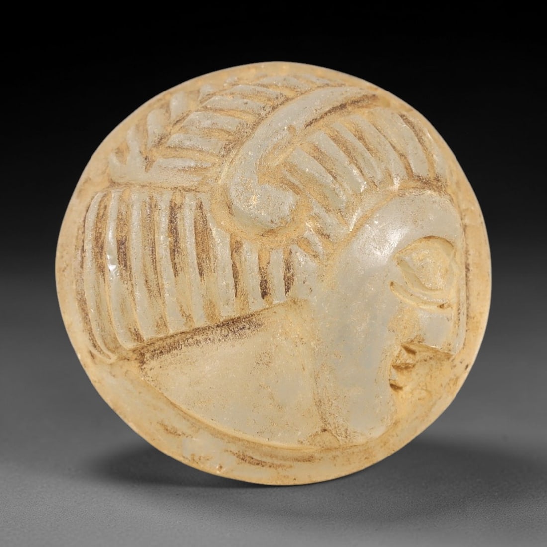 Ancient Glass Medieval Amulet - Near Eastern Relic, 89AL: This artifact is crafted from ancient glass, molded into a circular form and carved in relief with a profile human head, featuring detailed facial features and stylized hair with incised linear patter