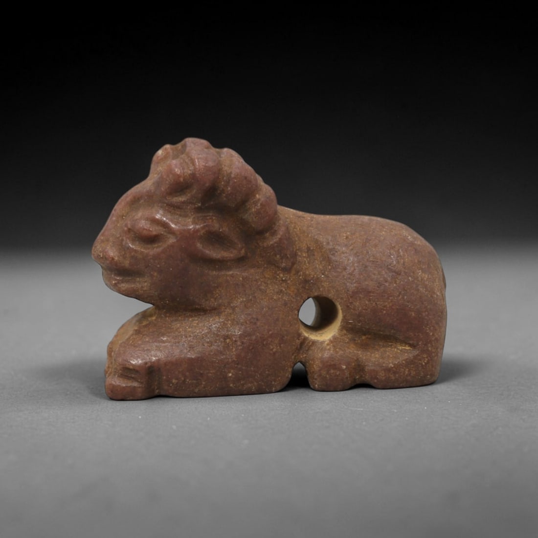 Ancient Stone Ram Amulet - Near Eastern Figurine, 91XV: This amulet is carved from a warm reddish-brown stone with natural earthy patina and subtle tonal variations across its surface, executed in a stylized manner that emphasizes the recumbent ram's form.