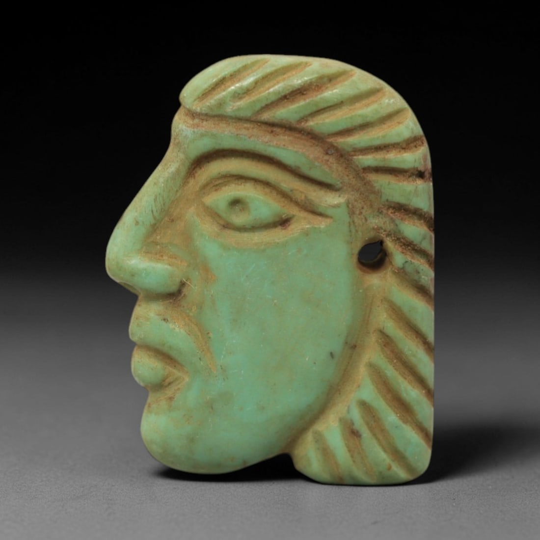 Ancient Turquoise Profile Head Amulet - Egyptian Figurine, 1XV: Carved from turquoise, this amulet depicts a stylized human head in profile, facing left, with incised linear details defining a textured headdress or hair, almond-shaped eyes, a prominent nose, and c