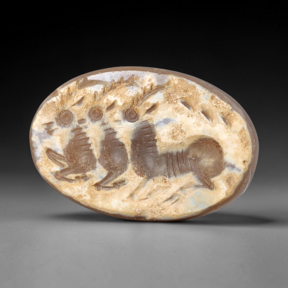 Ancient Agate Intaglio - Near Eastern Animal Amulet, 84AR: This intaglio is carved from banded agate, featuring a layered, cameo-style relief of three stylized animal figures in striding pose. The iconography depicts horned quadrupeds with incised linear deta