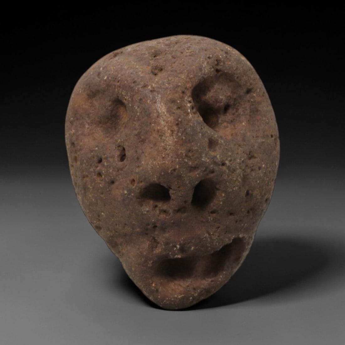 Ancient Stone Anthropomorphic Mask - African Tribal Amulet, 69FW: Carved from a porous, earthy-brown stone with a naturally pitted surface, this artifact takes the form of a stylized anthropomorphic mask with deeply recessed circular eye sockets, a double-nostril na