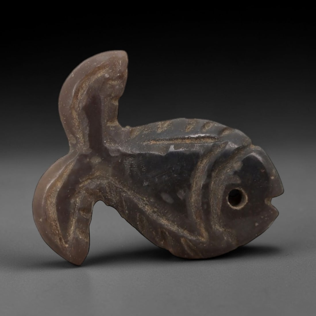 Ancient Stone Fish Amulet - Egyptian Carved Pendant, 84AG: This amulet is carved from a dark, translucent stone with a smooth, weathered patina and subtle surface wear. The form is a stylized fish, with incised linear details defining the dorsal, ventral, and