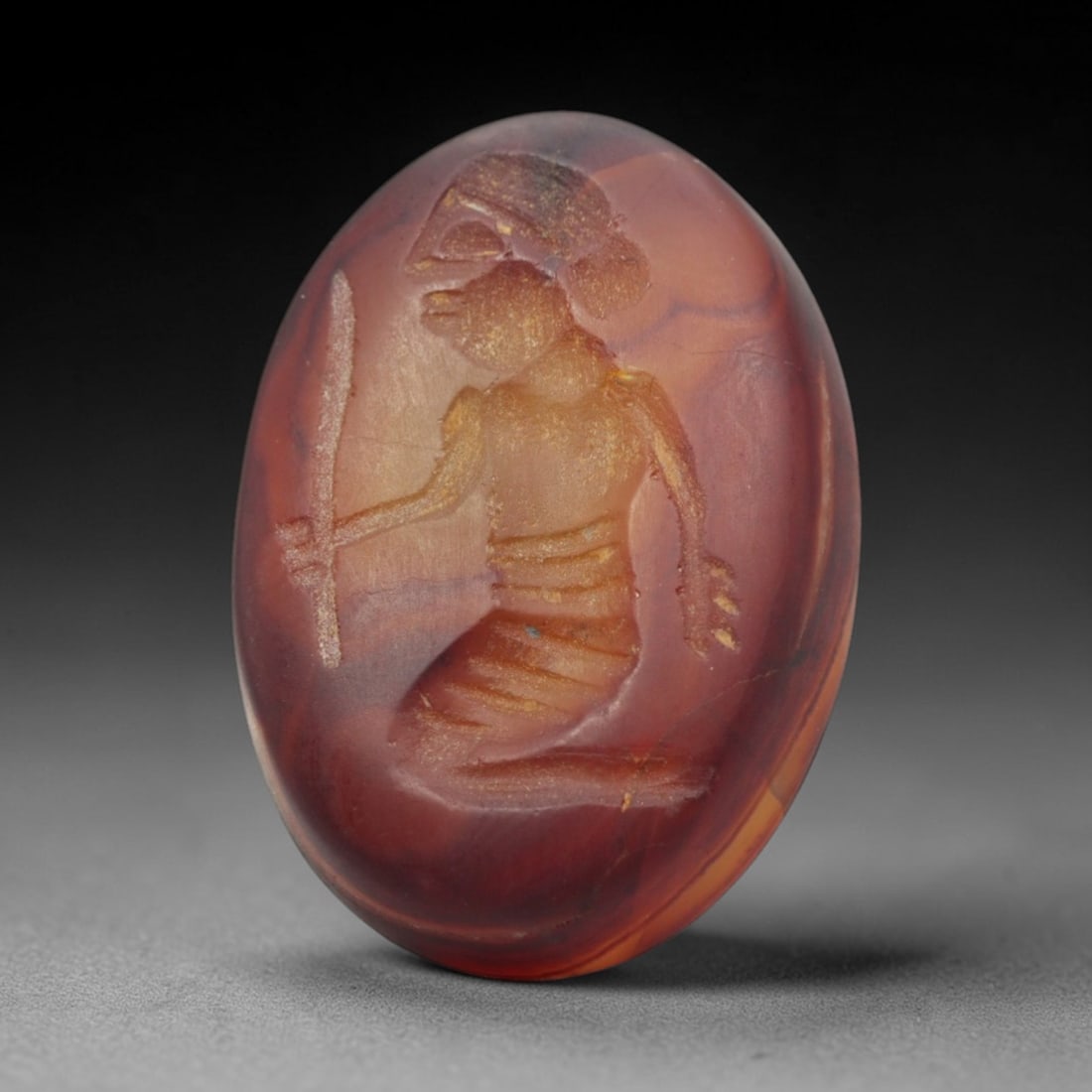 Ancient Agate Intaglio - Near Eastern Kneeling Warrior, 27ZW: Carved from rich banded agate, this oval intaglio depicts a kneeling warrior figure wielding a sword, with incised linear detailing to denote anatomical structure and decorative elements, characterist
