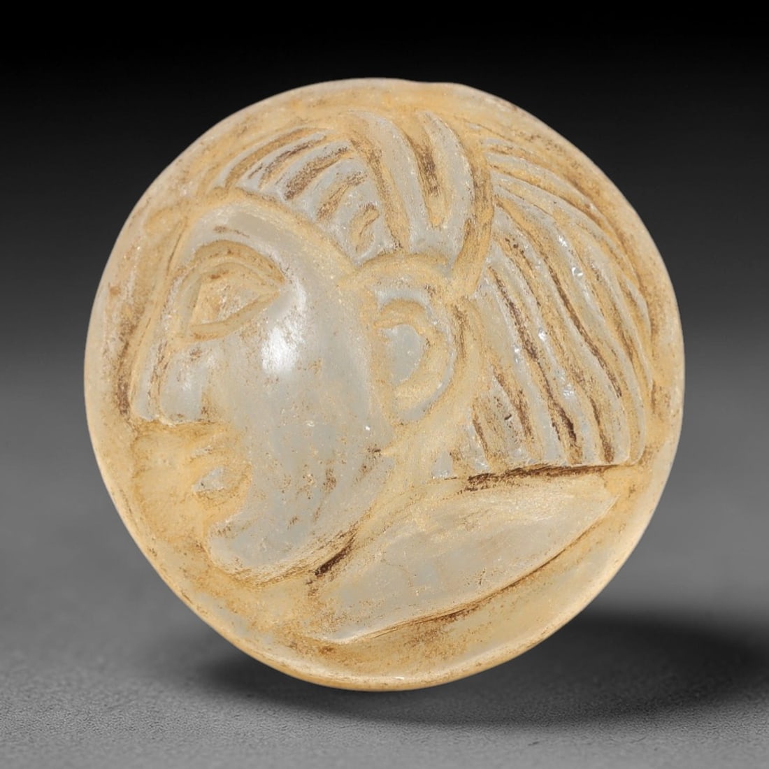Ancient Glass Medieval Amulet - Near Eastern Relic, 90AL: This artifact is crafted from ancient glass, formed into a circular disc and carved in relief with a profile human head, featuring stylized linear hair patterns, defined facial features, and a distinc