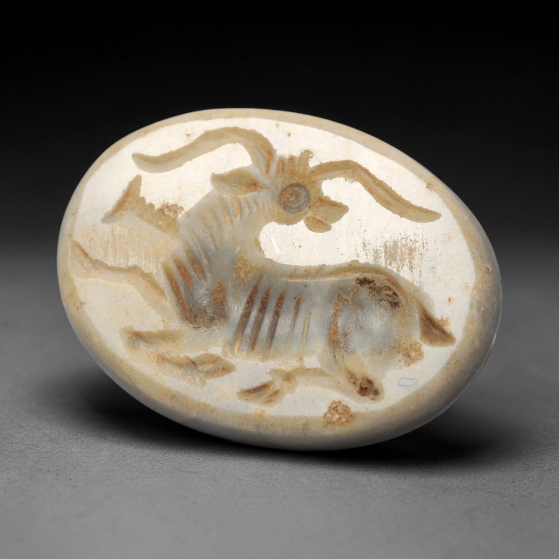 Ancient Agate Intaglio Bead - Near Eastern Ibex Motif, 38ZW: Carved from pale banded agate, this oval intaglio bead features a recessed depiction of a recumbent ibex with prominent curved horns, incised linear detailing to denote anatomical structure and patter