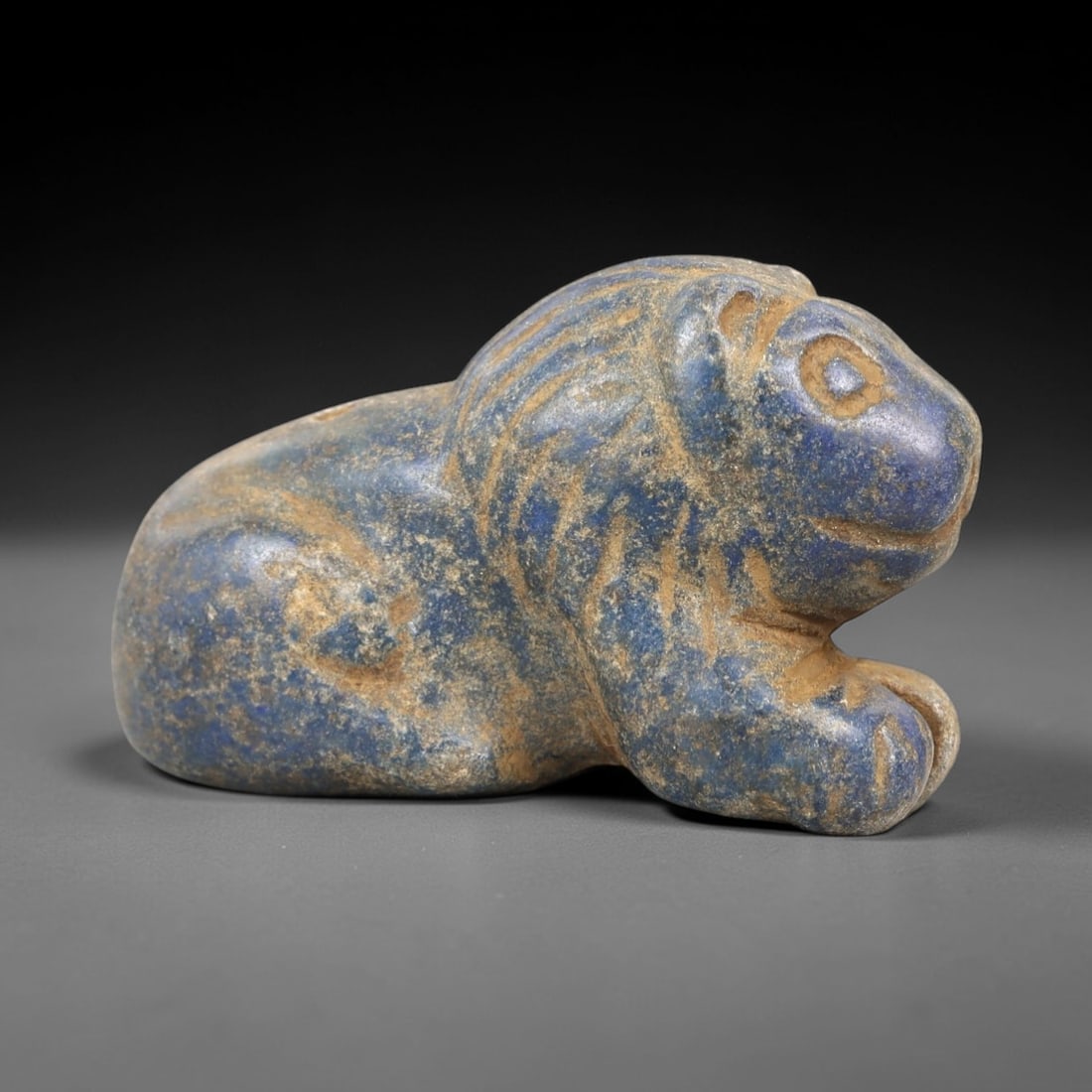 Ancient Lapis Lazuli Feline Amulet - Near Eastern Lion Pendant, 30AK: Carved from deep blue lapis lazuli, this reclining feline amulet depicts a stylized lion with incised linear patterns defining its mane, facial features, and paw details. A subtle perforation near the