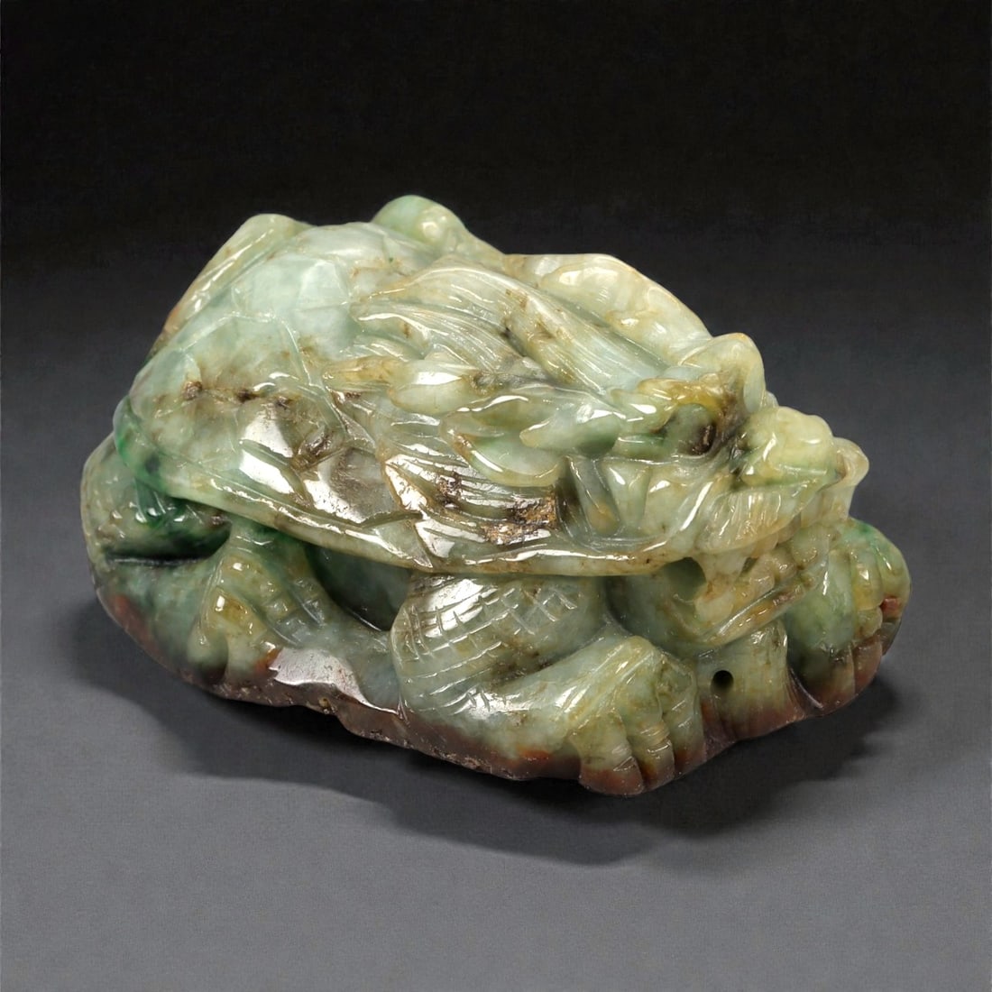 A Chinese Jadeite Jade Bixi Figure, 20th Century, 70FB: Carved from mottled jadeite jade displaying pale green tones with darker inclusions and areas of brownish surface staining, the figure is worked in the round and modeled as a bixi, the mythological cr