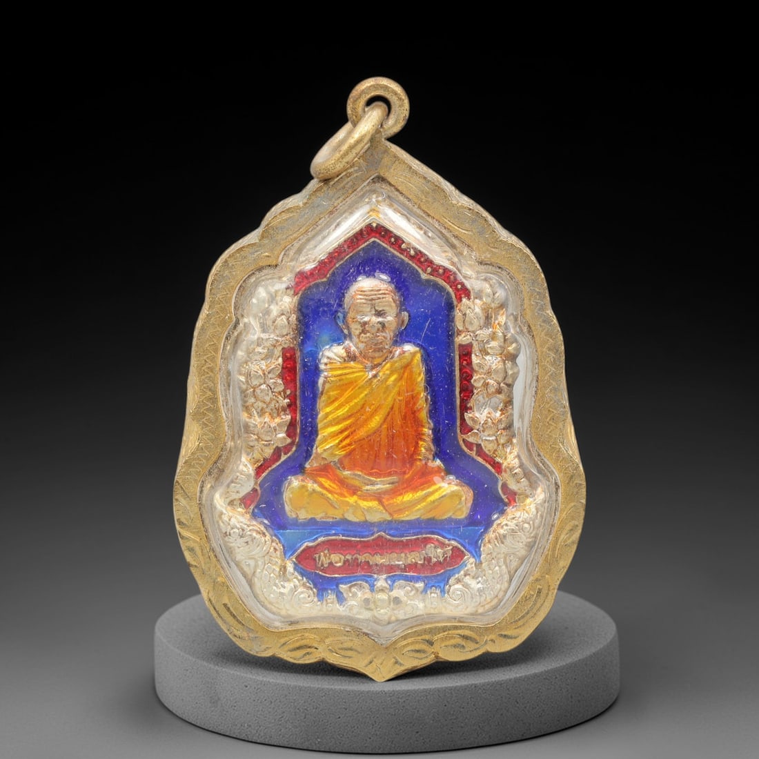 A Thai Silver Buddhist Amulet in Gold Frame, Late 20th Century, 100FP: A shield-shaped silver amulet, measuring 5 x 3.5 cm, set within an ornate gold frame with a suspension loop. The obverse depicts a seated Buddhist monk in meditative posture, rendered in gold and oran