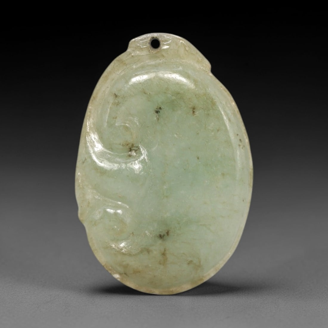 A Chinese Jadeite Jade Pendant, 20th Century, 84FR: Carved from pale green jadeite jade with natural dark inclusions and faint tonal variations, the pendant has an oval form with a drilled suspension hole at the apex. A simplified swirling relief motif