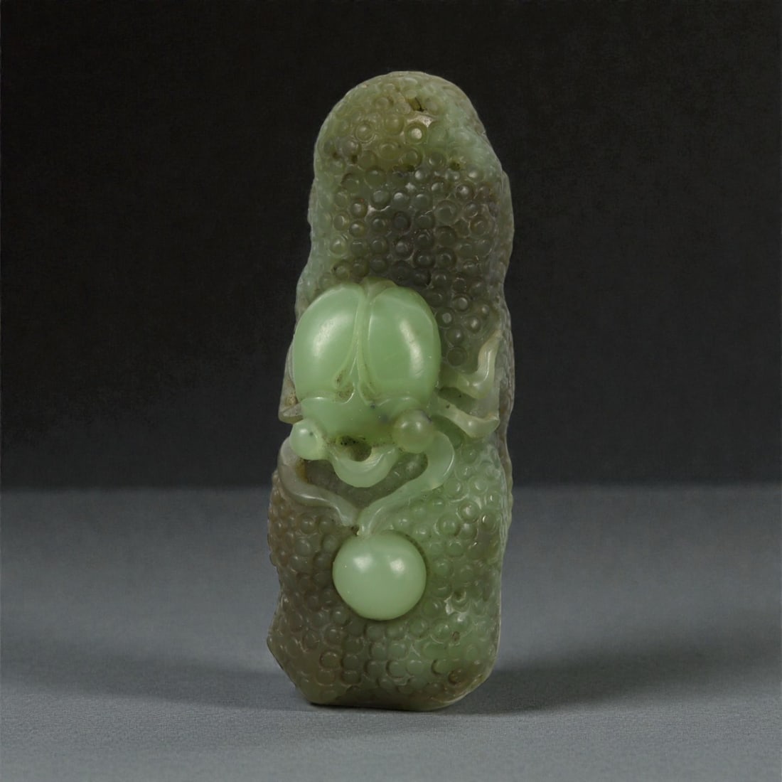 A Chinese Nephrite Jade Spider-and-Fruit Pendant, 20th Century, 12FB: Carved from green nephrite jade with darker mottling and areas of translucency, the pendant is modeled as an elongated fruit with a densely textured surface composed of rounded nodules. The front is c