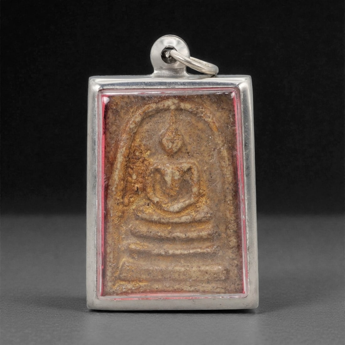 A Thai Pressed Powder Buddha Amulet in Metal Case, 20th Century, 44ZH: The amulet is formed from pressed powder with residual gilding, depicting a seated Buddha in bhumisparsha mudra, enclosed within a rectangular metal case with a red resin border. The reverse of the am