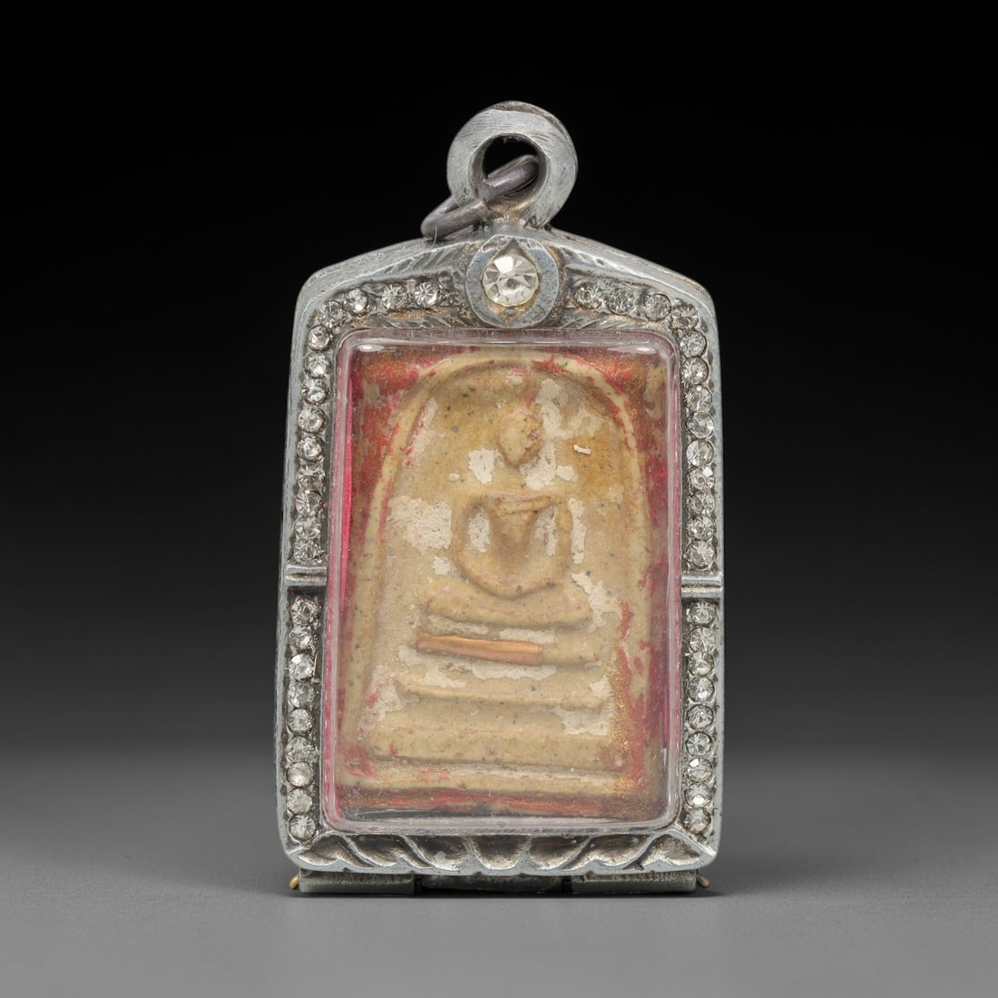 A Thai Terracotta Buddha Amulet in Silver Case, 20th Century, 72ZH: The amulet is formed from terracotta, with a light beige base and embedded particulate matter visible on the reverse. The obverse depicts a seated Buddha in bhumisparsha mudra, with the figure seated