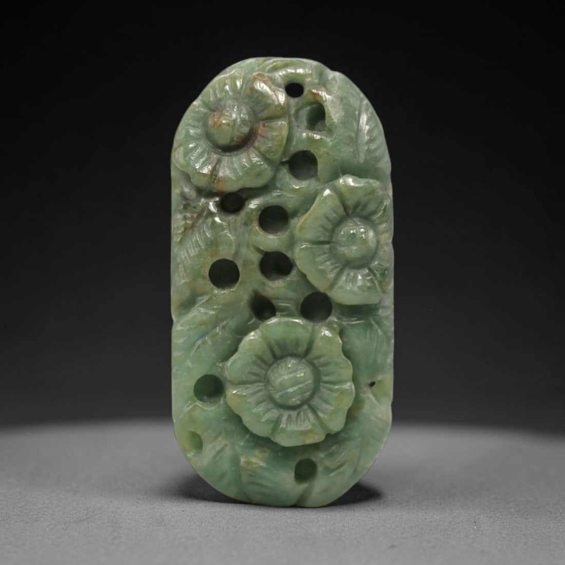 A Chinese Jadeite Jade Floral Pendant, 20th Century, 52FR: Carved from light green jadeite jade with natural tonal variation and minor inclusions, the pendant is executed in openwork relief. Three floral forms with radiating petals and central bosses are arra