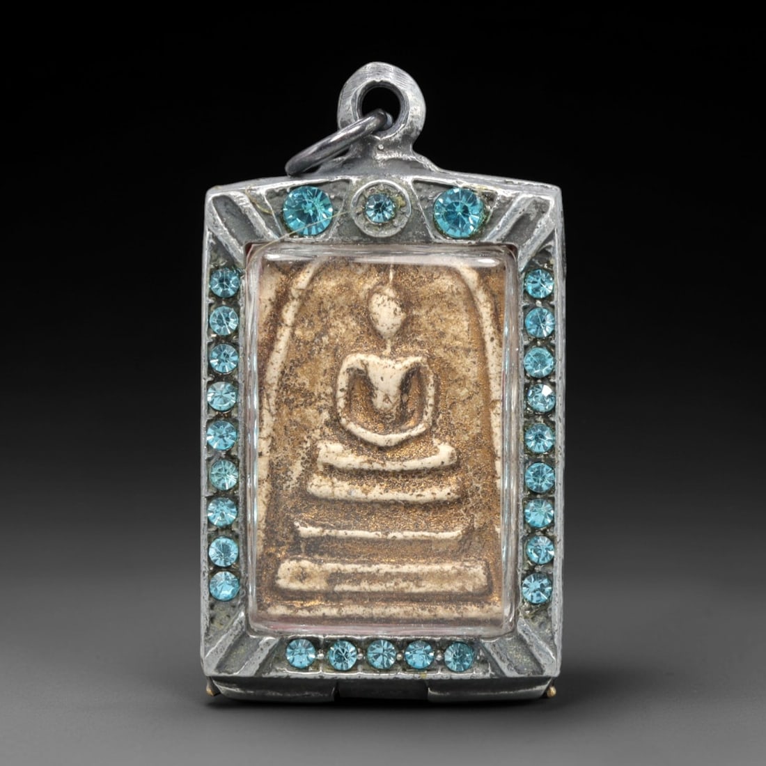 A Thai Phra Somdet Clay Amulet in a Gem-Set Silver-Tone Case, 20th Century, 24ZK: This rectangular Phra Somdet amulet is crafted from sacred beige clay and encased in an ornate silver-tone metal case with a suspension loop at the apex. The obverse of the case is elaborately set wit