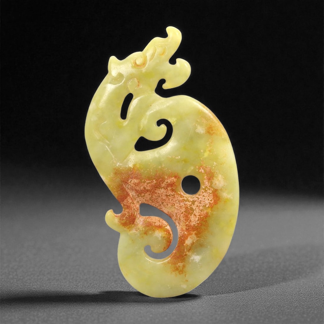 A Chinese Archaistic-Style Hetian Jade Dragon Plaque, 20th Century, 73FH: Carved from pale yellow-green Hetian jade with areas of pronounced russet and iron-oxide staining, the plaque is formed as a stylized dragon in profile and executed in openwork. The body is elongated