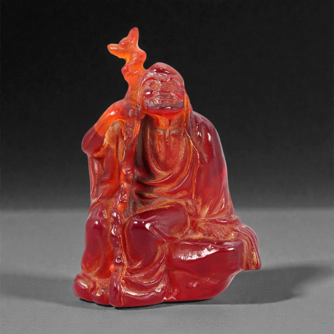 A Chinese Amber-Resin Figure of a Luohan, 20th Century, 44FH: Cast or carved from translucent amber-colored red resin with internal tonal variation, localized inclusions, and minor entrapped bubbles, the figure represents a seated luohan shown resting against a