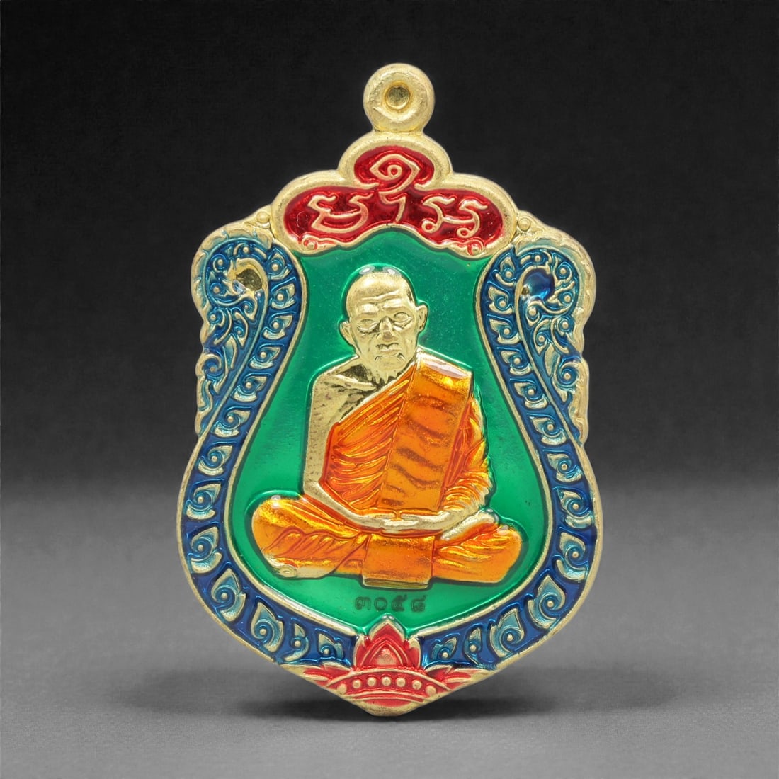 A Thai Gilt-Metal and Enamel Monk Portrait Amulet, 21st Century, 69FP: Cast from metal with a gilt surface finish, the amulet has a shield-shaped form with an integral suspension loop at the apex. The obverse features a high-relief gilt bust portrait of a seated Buddhist