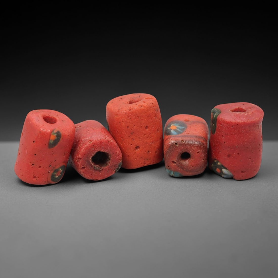 A Group of Venetian Red Mosaic Glass Beads, 19th-20th Century, 25ZM: Fabricated from opaque red glass, the group consists of five cylindrical beads with central perforations for suspension. Two beads display embedded circular mosaic inlays of green, orange, and white;