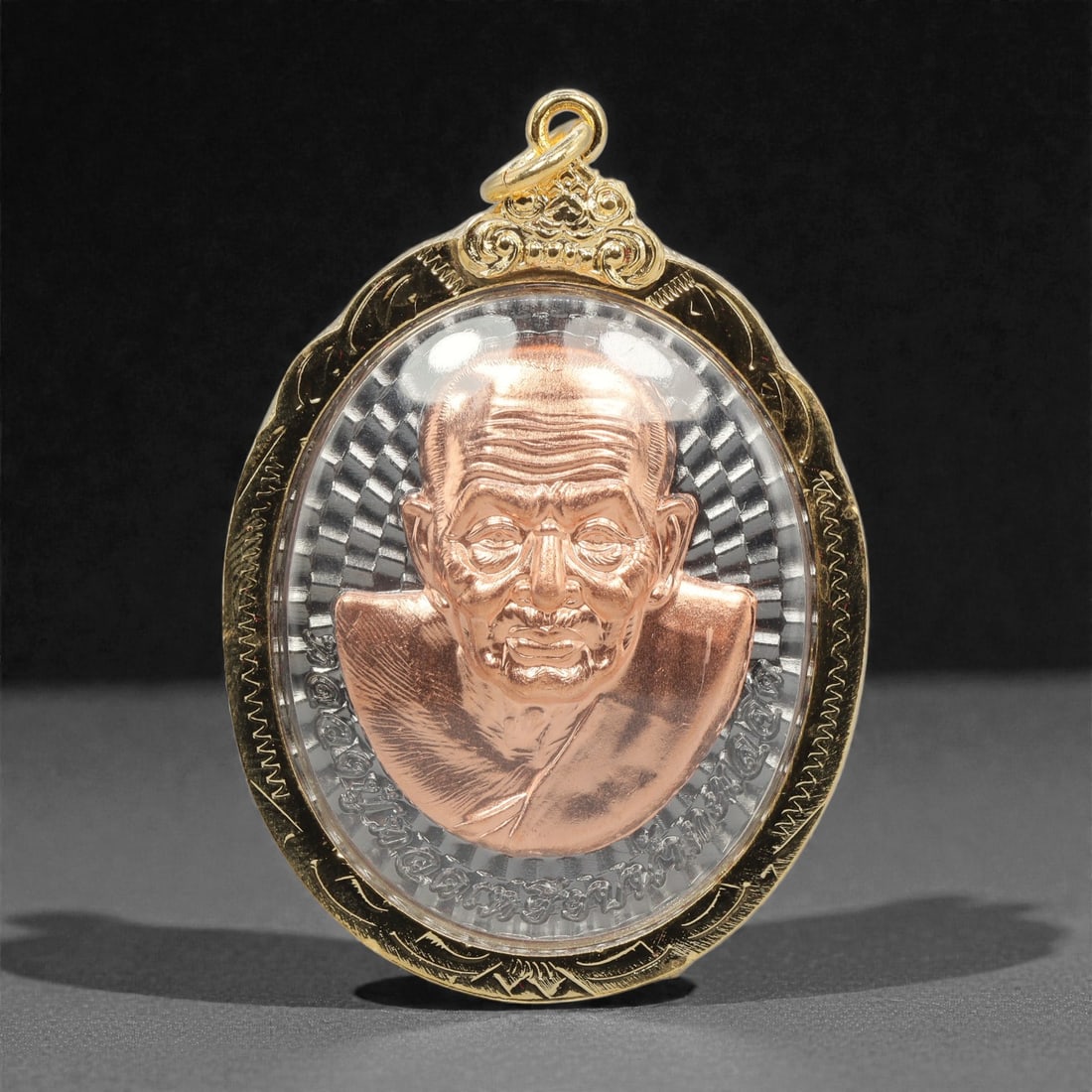 A Thai Bi-Metallic Monk Portrait Amulet with Gilt-Metal Frame, 21st Century, 64FP: The amulet is of oval form, featuring a high-relief copper-toned bust portrait of a Buddhist monk on the obverse, set against a silver-toned radiating linear ground with incised Thai script arranged i