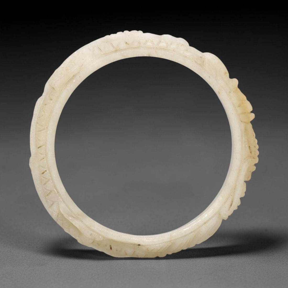 A Chinese Jadeite Jade Bangle Bracelet, 20th Century, 38FS: Carved from pale white jadeite jade with subtle natural inclusions and minor surface discoloration, the bangle is formed as a continuous circular band. The exterior surface is decorated with raised re