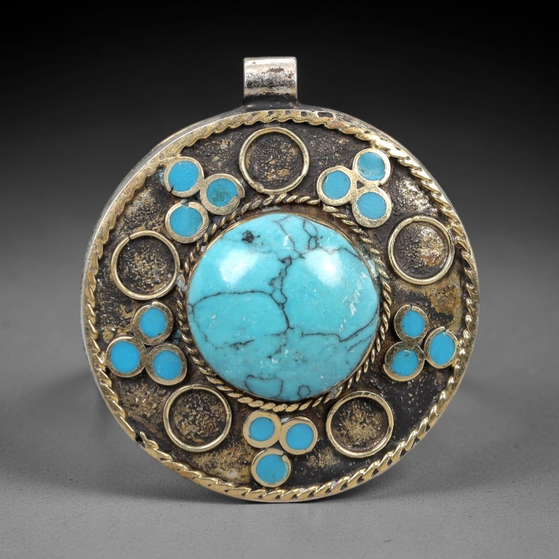 A Tibetan Silver Pendant with Turquoise Inlay, 20th Century, 95XM: Formed as a circular silver plaque, the pendant is centered by a large, polished turquoise cabochon with natural dark veining. The cabochon is set within a bezel of coiled silver wire, surrounded by m